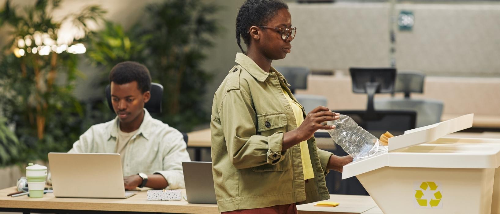 office setting, woman in foreground putting plastic bottle in recycling, man in background at desk on computer