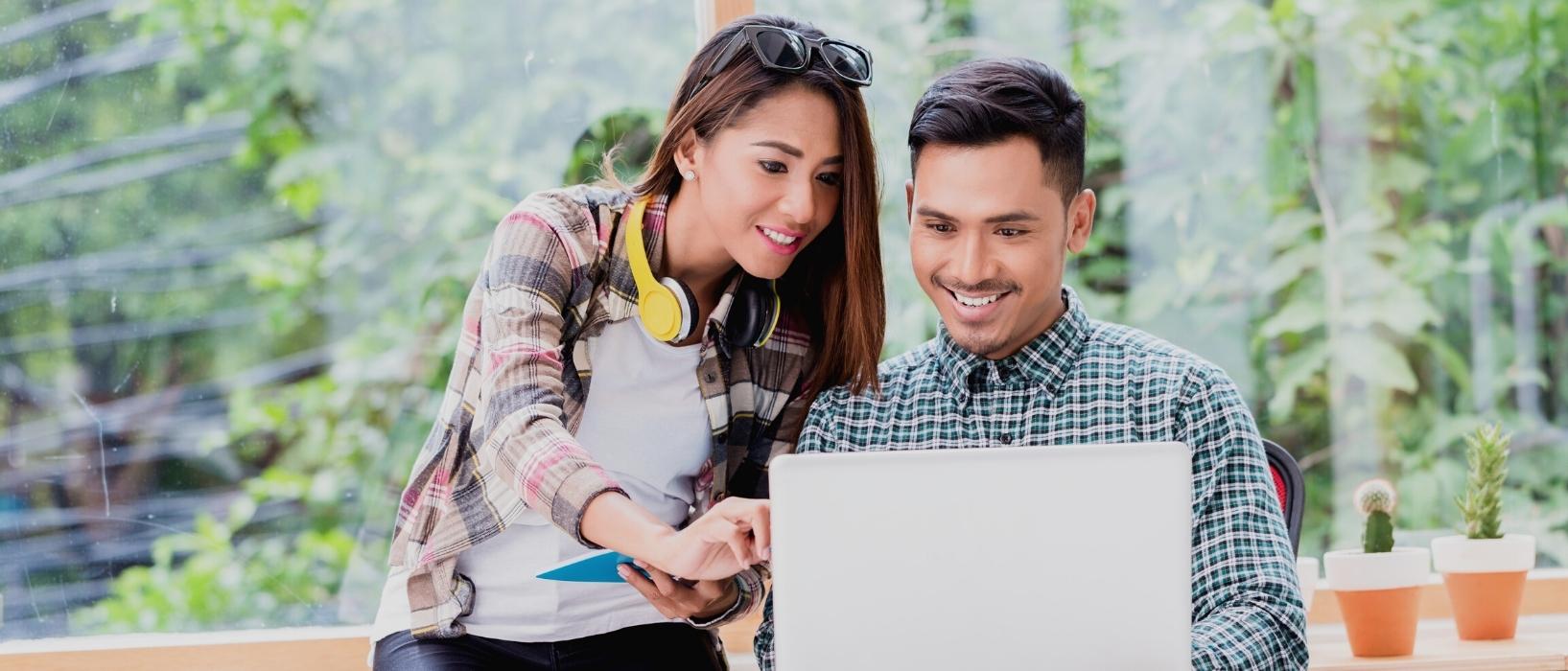 man in shirt looking for job on laptop, woman with headphone around neck leaning over and helping him