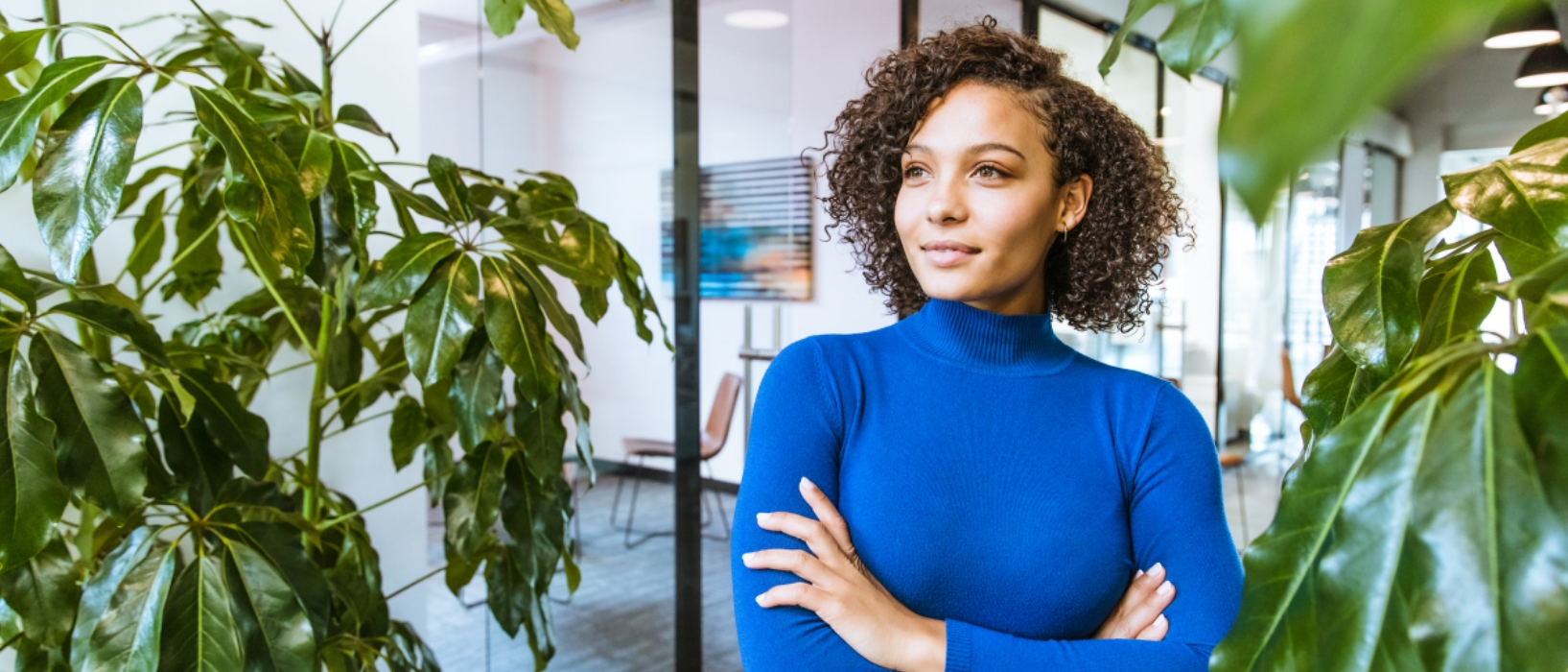 woman from torso up standing with arms crossed in blue jumper in middle of office surrounded by plants