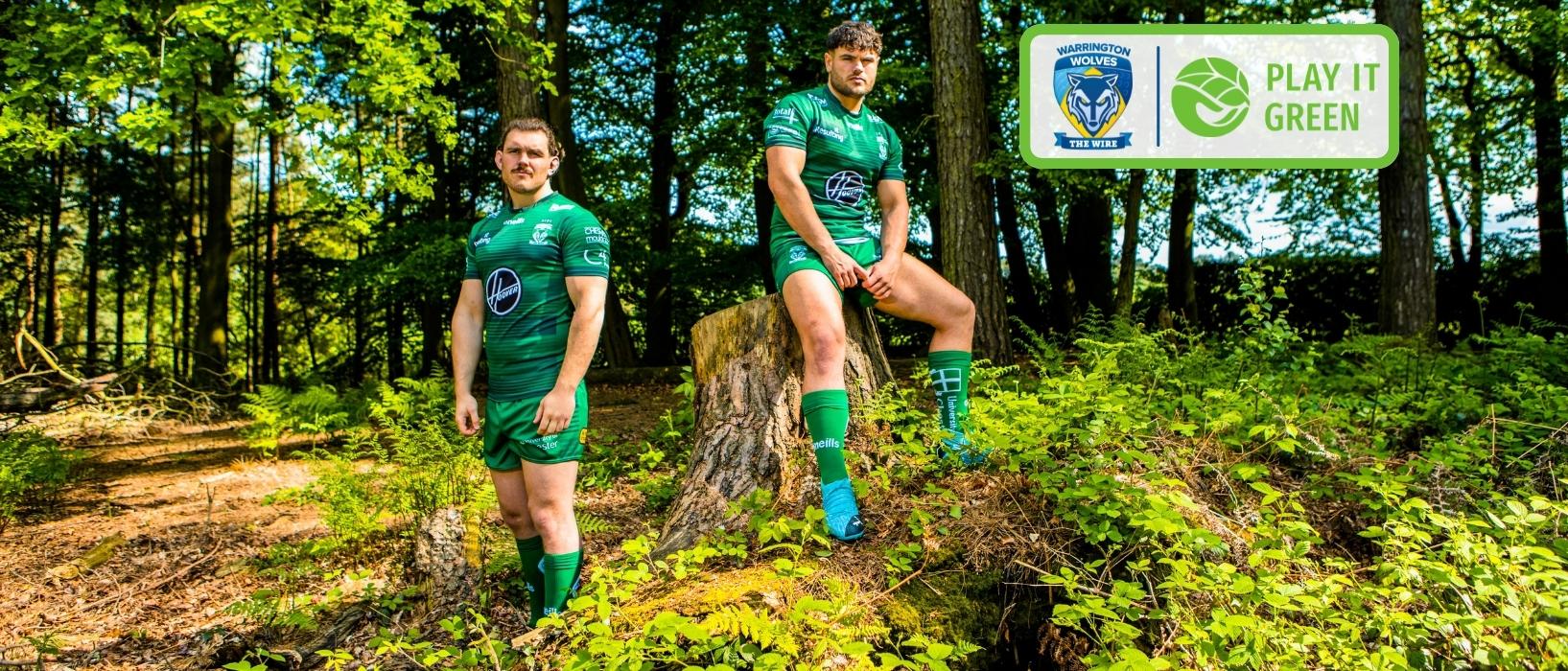 two warrington wolves players in full green rugby kit in forest, one is standing while the other sits above on a tree stump