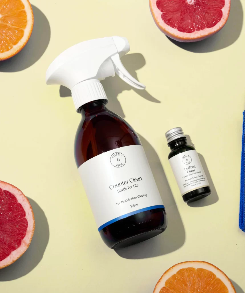 glass spray cleaning bottle with white lid and label with Purdy and Figg logo, smaller glass bottle with silver cap beside it, both lying on yellow background with orange and red citrus fruits cut open around border