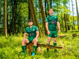 warrington wolves players in full rugby kit stand in forest, one on the left sits on a tree stump in front of the on standing up behind him