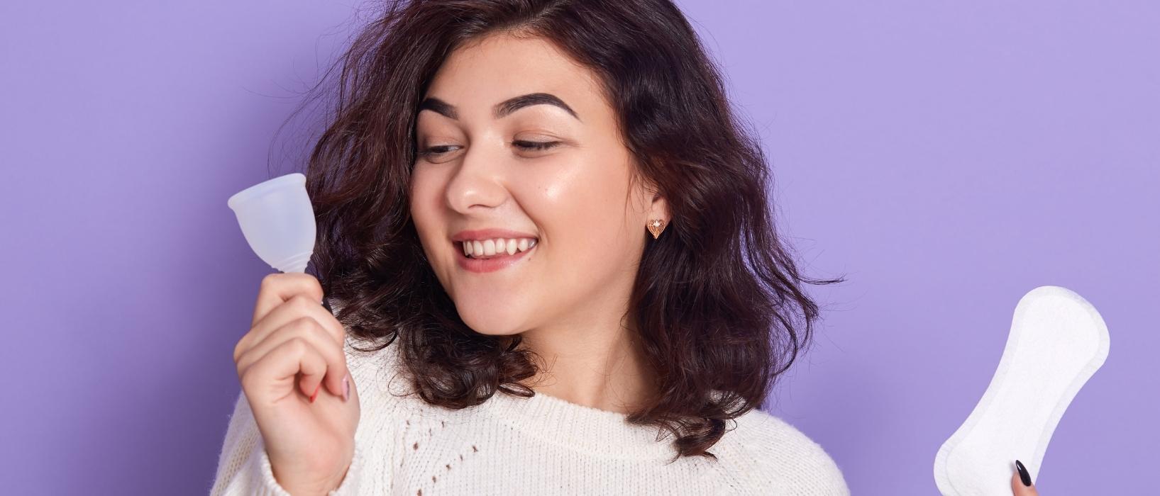 dark-haired woman shown shoulders up against purple background, holding pad in one hand and menstrual cup in another, she is smiling at menstrual cup